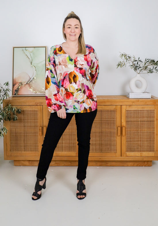 Woman wearing a colorful floral top and black pants standing in a room with wooden furniture and plants.
