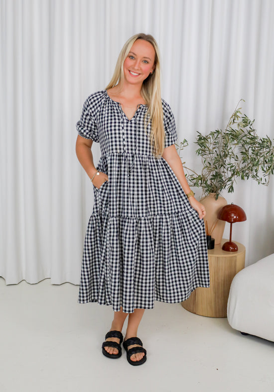 Woman wearing a black and white checkered dress in a room with a white curtain and plant.