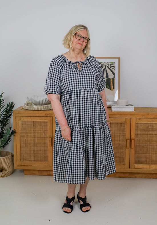 Woman wearing a black and white checkered dress standing in a room with wooden furniture and a plant.