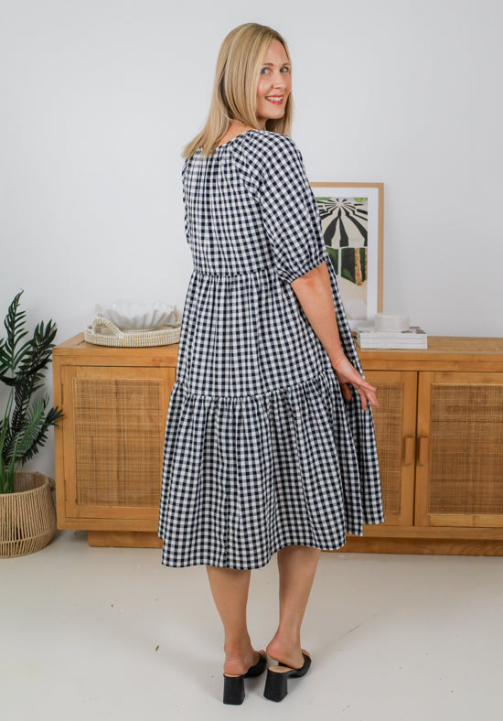 Woman wearing a black and white checkered dress standing in a room with wooden furniture and a plant.