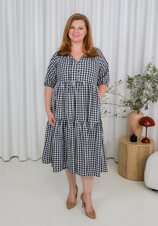 Woman wearing a black and white checkered dress standing in a room with a curtain and plant in the background.
