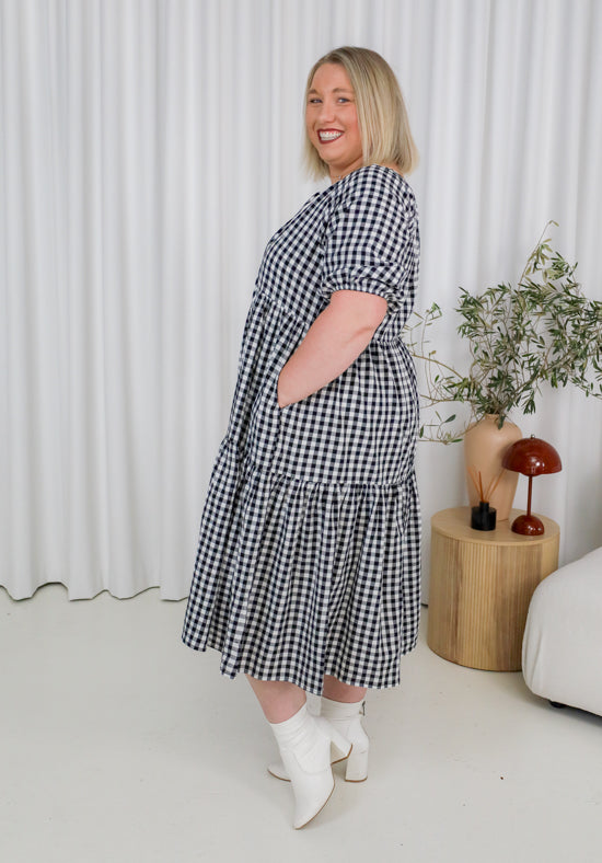 Woman wearing a black and white checkered dress standing in a room with a white curtain and a small table with a plant and lamp.