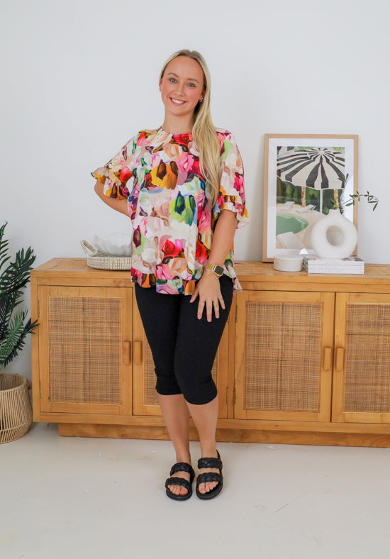 Woman wearing a colorful floral blouse and black capri pants standing in a room with a wooden cabinet and decorative items.