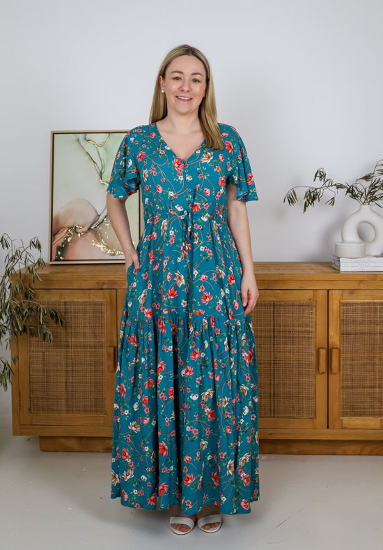 Woman wearing a teal floral dress standing in a room with wooden furniture and plants.