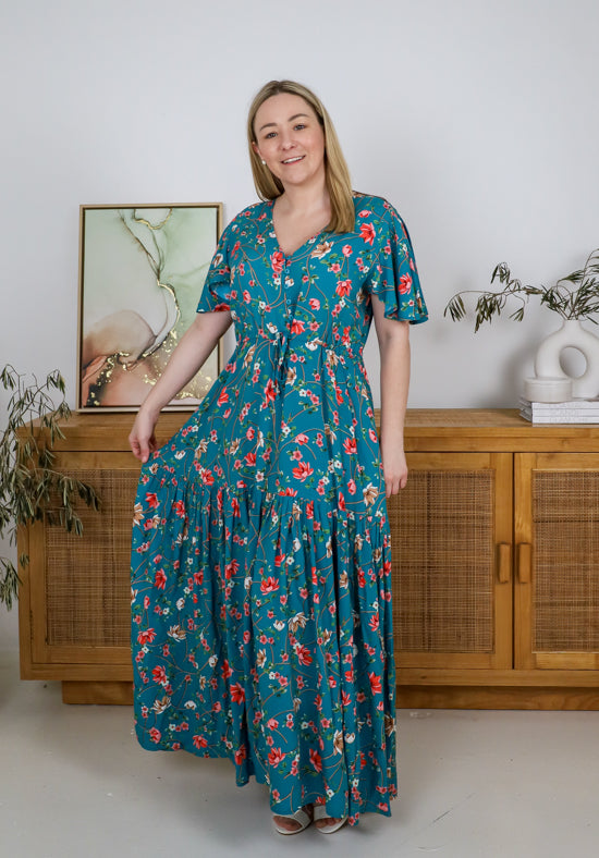 Woman wearing a teal floral dress standing in a room with wooden furniture and plants.