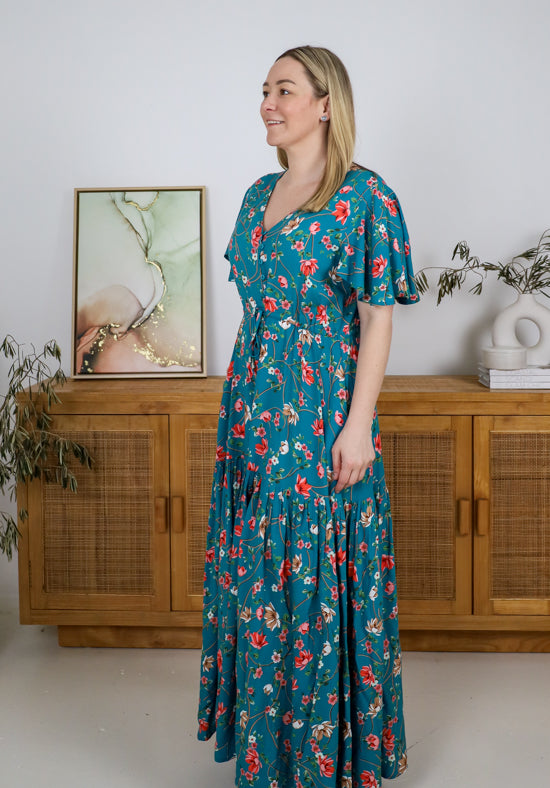 Woman wearing a teal floral dress standing in a room with wooden furniture and plants.