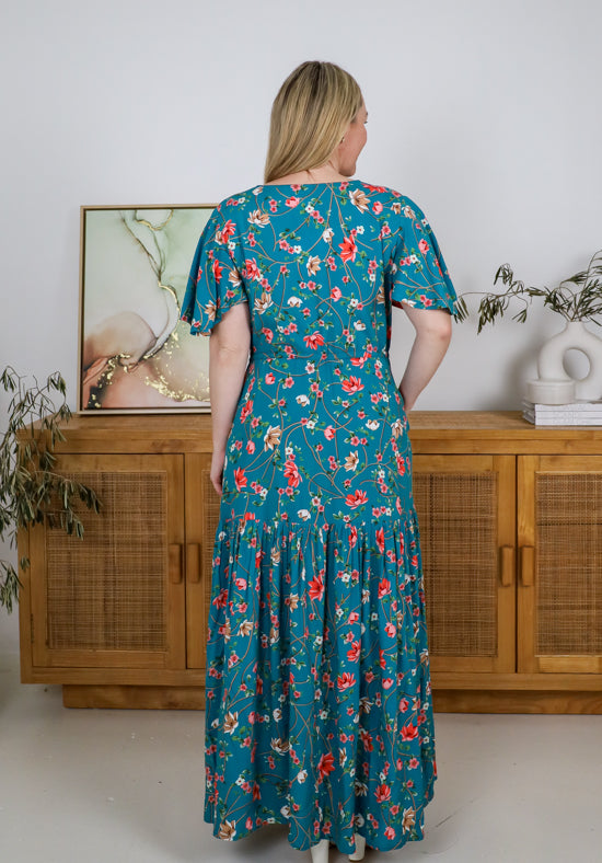 Woman wearing a floral dress standing in a room with wooden furniture and decor.