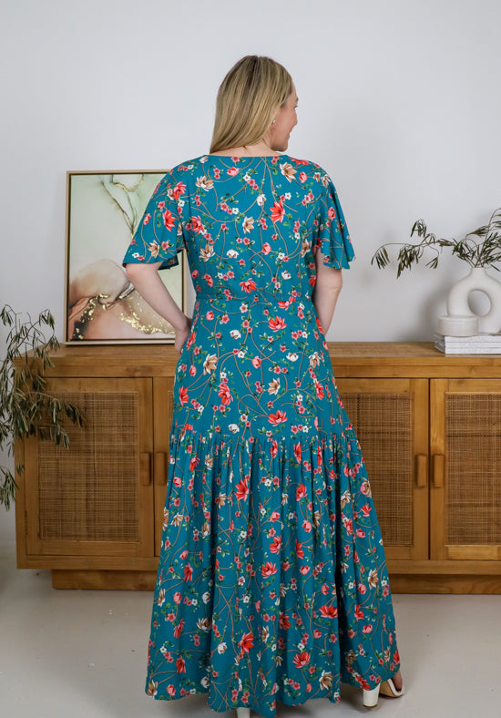 Woman wearing a teal floral dress standing in a room with wooden furniture and plants.