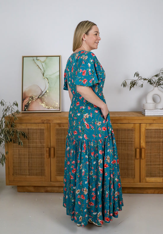 Woman wearing a teal floral dress standing in a room with wooden furniture and a painting.