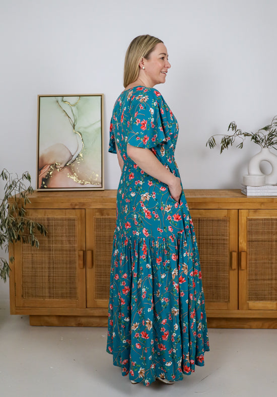 Woman wearing a teal floral dress standing in a room with wooden furniture and a painting.