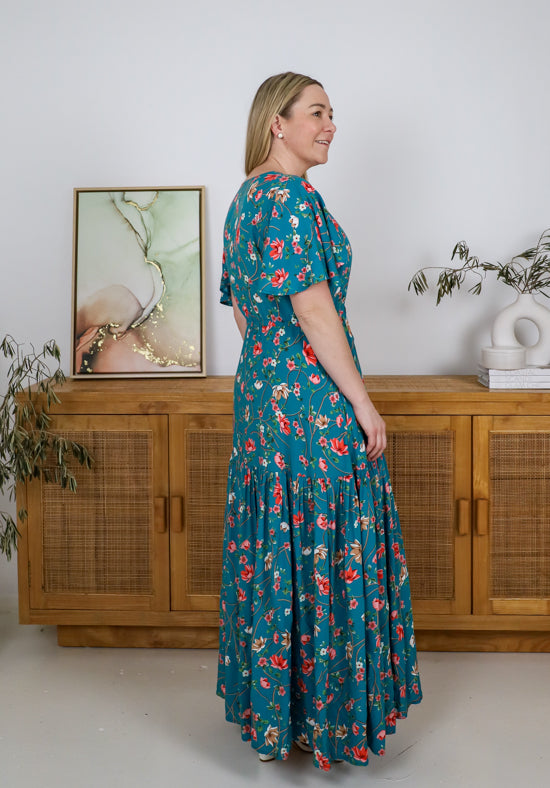 Woman wearing a teal floral dress standing in a room with wooden furniture and a painting on the wall.