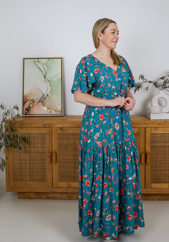 Woman wearing a teal floral dress standing in a room with wooden furniture and plants.