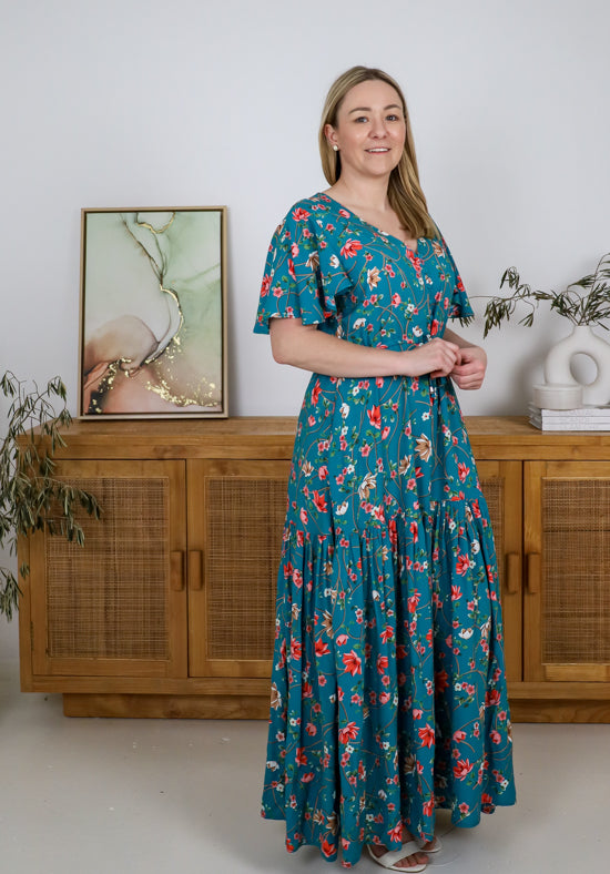 Woman wearing a floral dress standing in a room with wooden furniture and plants.