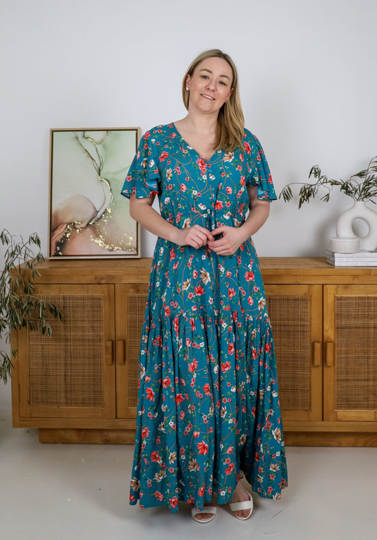 Woman wearing a teal floral dress standing in a room with wooden furniture and decorative items.