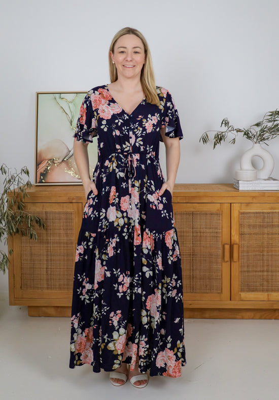 Woman wearing a floral dress standing in a room with wooden cabinets and plants.