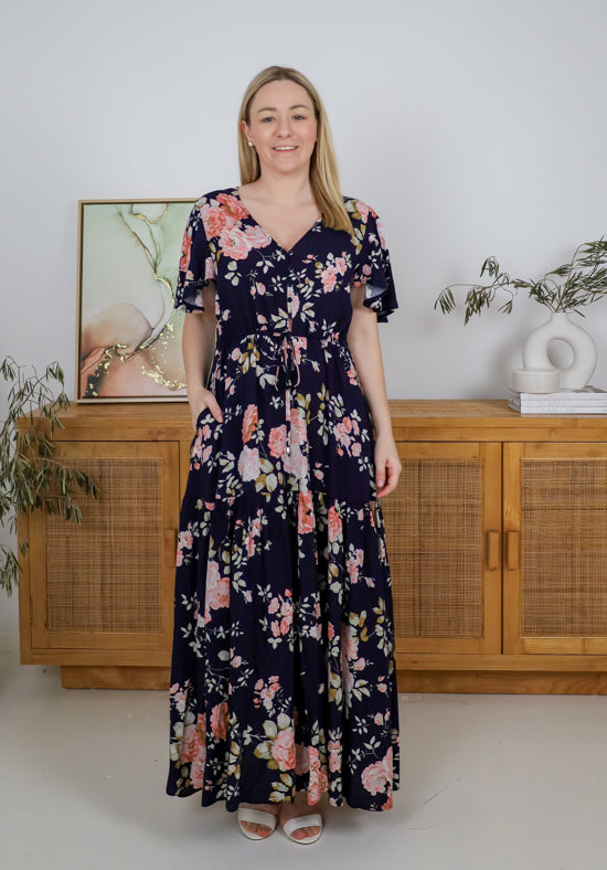 Woman wearing a floral dress standing in a room with wooden furniture and plants.