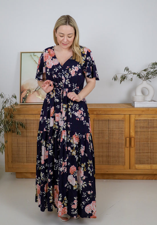 Woman wearing a navy floral dress standing in a room with wooden furniture and plants.