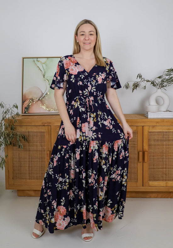 Woman wearing a floral dress standing in a room with wooden furniture and plants.