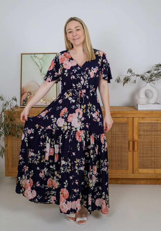 Woman wearing a floral dress standing in a room with a wooden cabinet and plants.