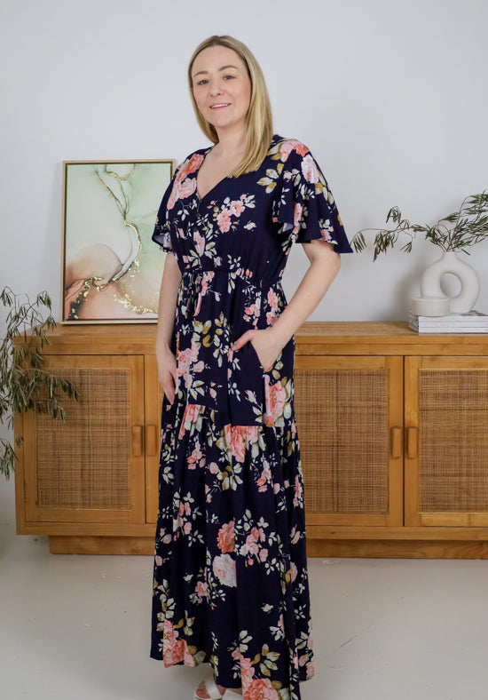 Woman wearing a floral dress standing in a room with wooden cabinets and decorative items.