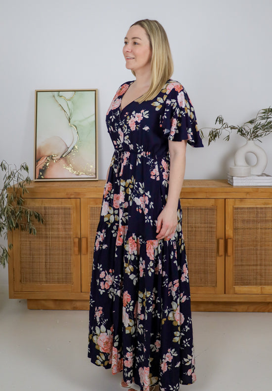 Woman wearing a floral dress standing in a room with wooden furniture and plants.