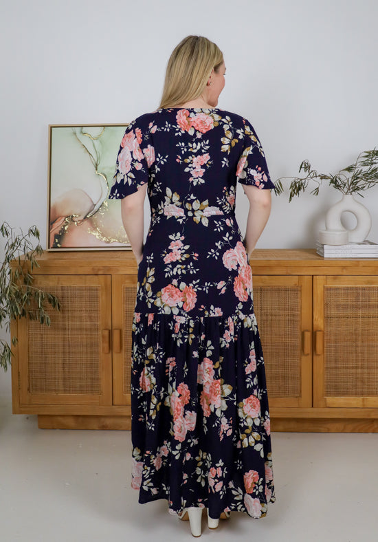 Woman wearing a floral dress standing in a room with wooden cabinets and decorative items.