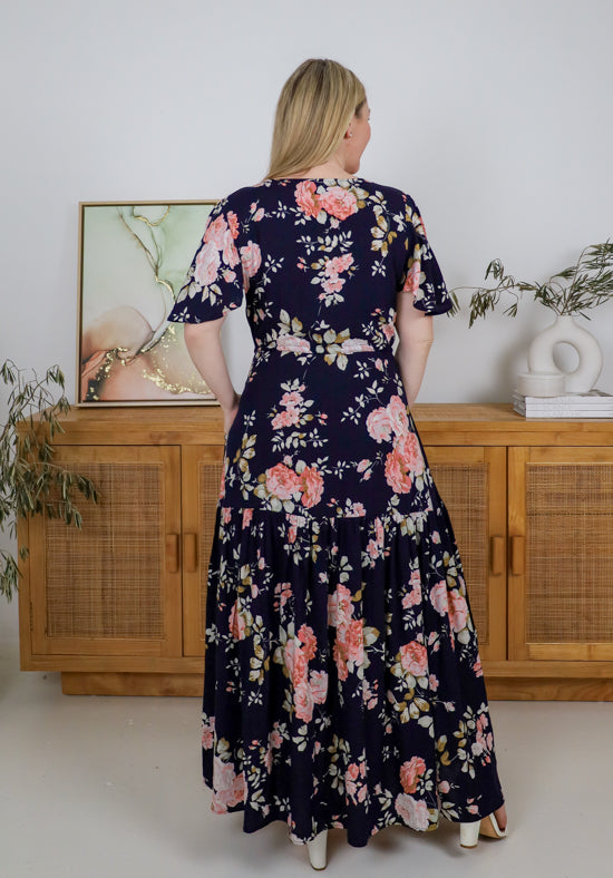 Woman wearing a floral dress standing in a room with wooden cabinets and decorative plants.