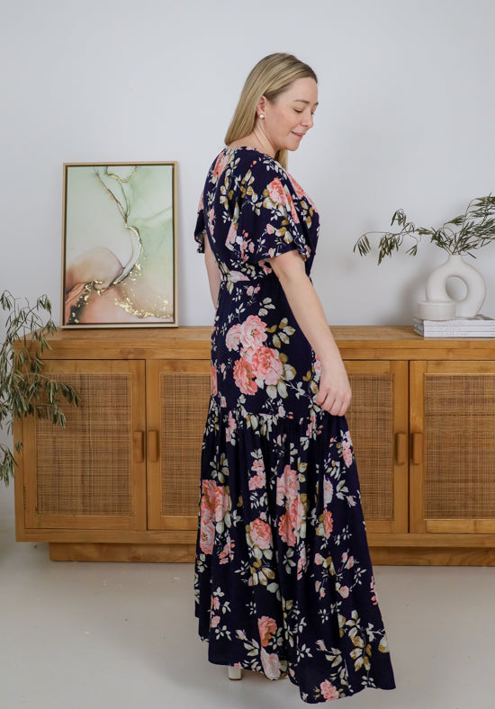 Woman wearing a floral dress standing in a room with wooden cabinets and plants.