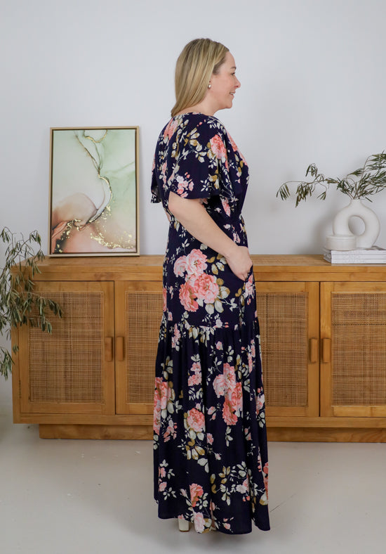 Woman wearing a floral dress standing in a room with wooden furniture and plants.